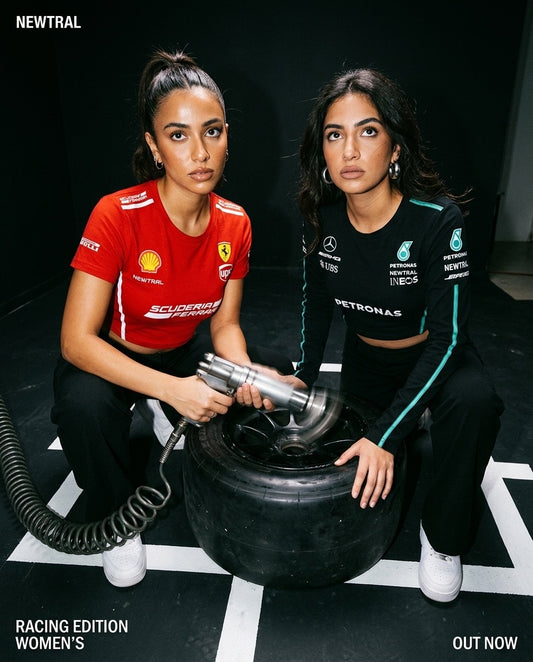 Ferrari Scuderia Women's Crop Tee (Red) and Mercedes-AMG Petronas Women's Longsleeve Crop (Black) bundle: Both models crouching around an F1 wheel with impact wrench, both garments fully visible, pit-lane floor markings